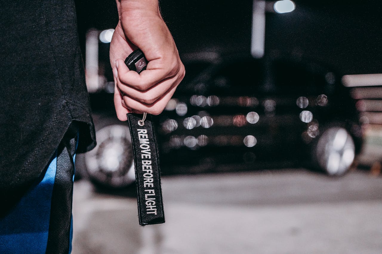 A hand holding a 'Remove Before Flight' keyring in front of a blurred car at night.